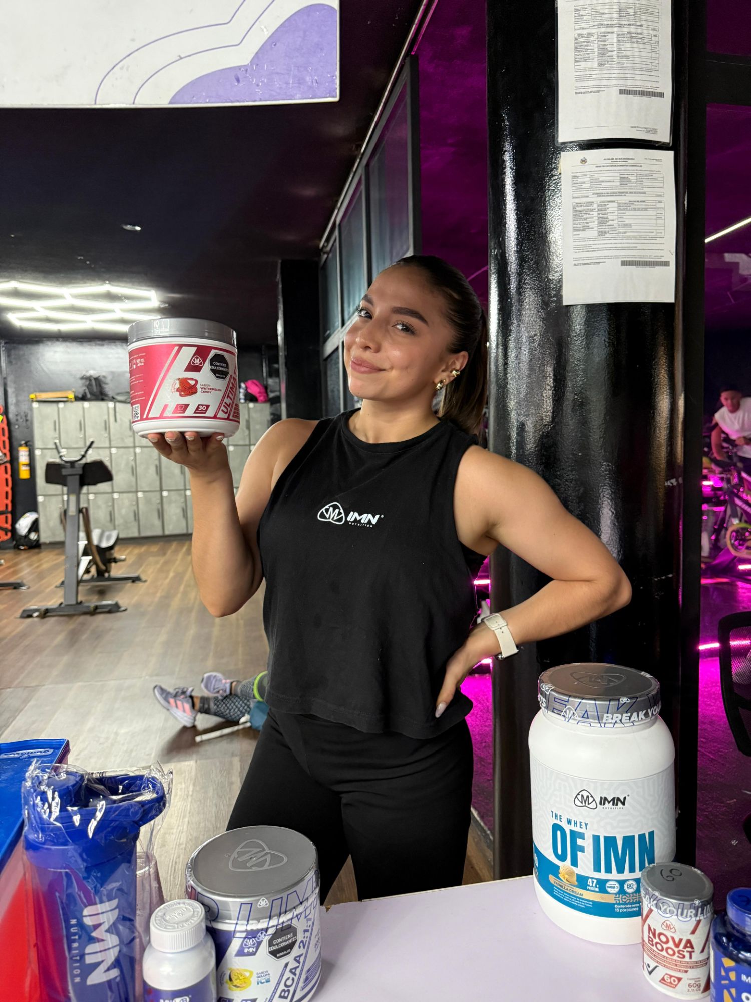Promotora IMN sonriendo en un gimnasio mientras muestra un Ultimate sabor watermelon candy