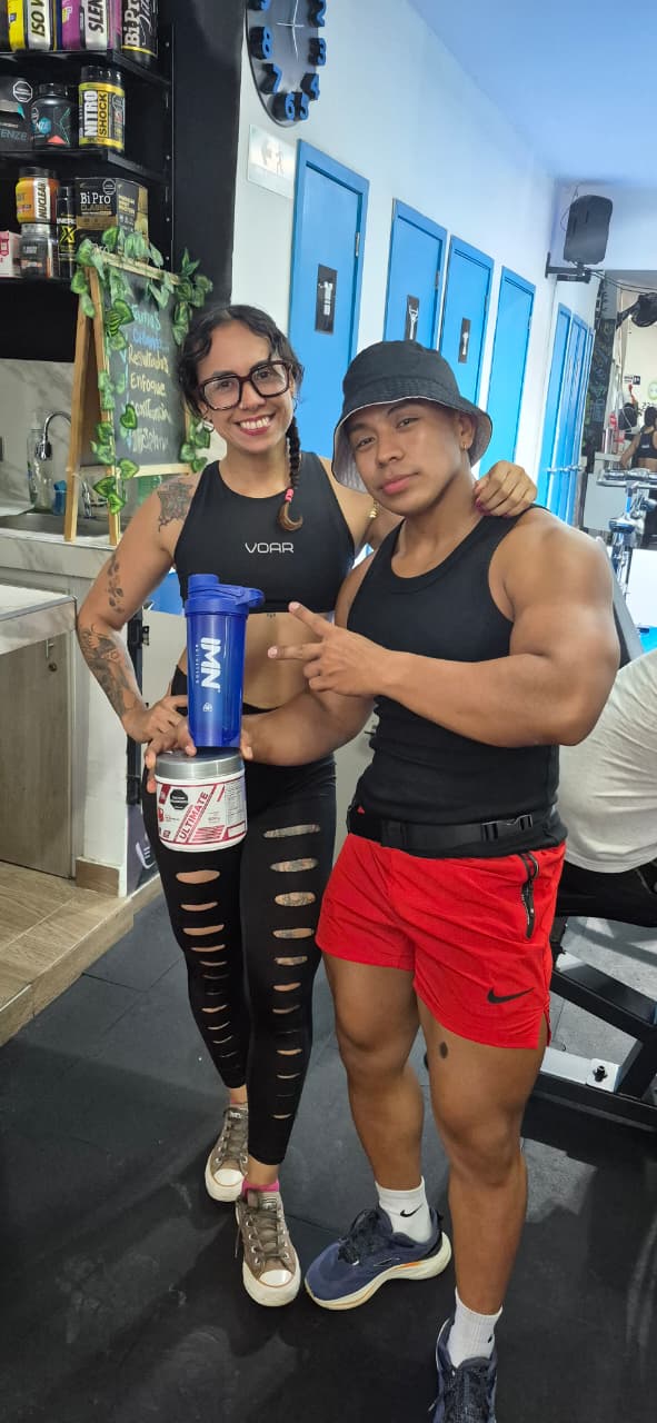 Promotora IMN junto a un cliente en un gimnasio; el cliente sostiene el producto que acaba de comprar y un shaker IMN