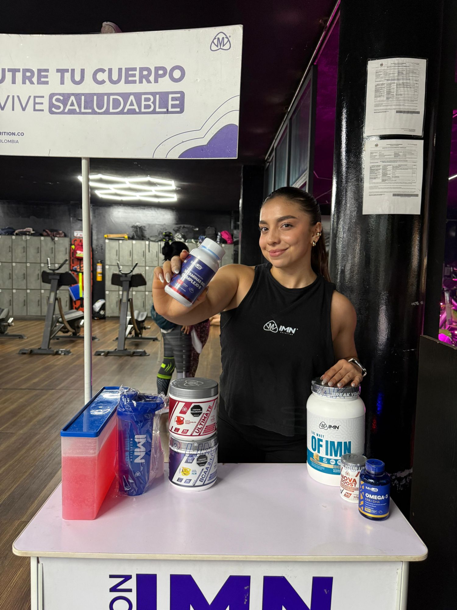 Promotora IMN con camiseta de la marca en un punto de impulso dentro de un gimnasio, mostrando un frasco de Complejo B junto a Ultimate, BCAA, The Whey, Nova Boost y Omega-3