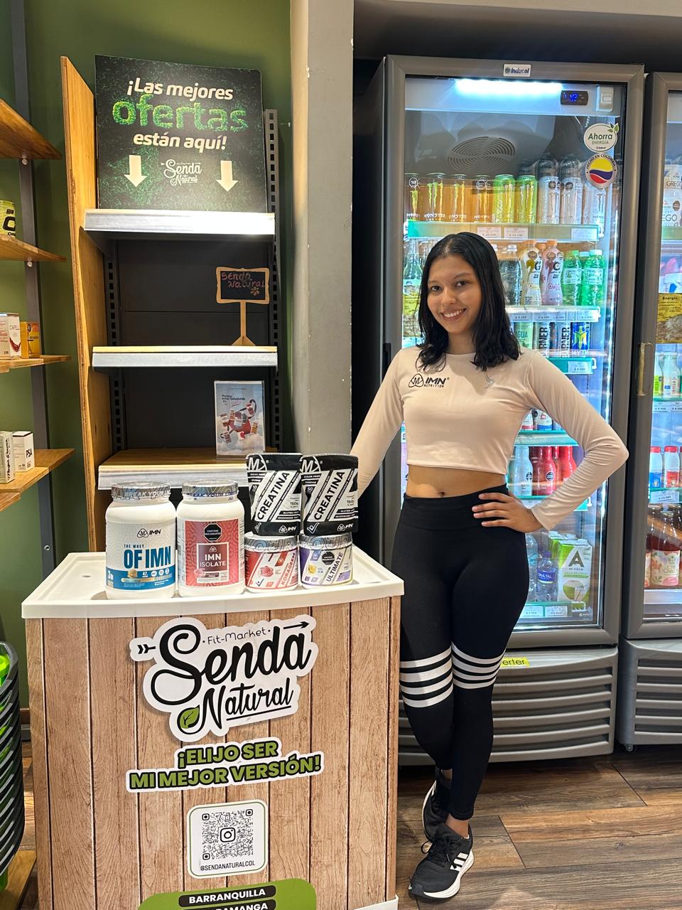 Promotora IMN de pie junto al exhibidor de una tienda saludable, con cabello recogido, uniforme IMN y postura abierta
