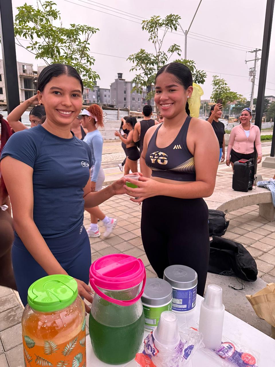 Promotora IMN ofreciendo una degustación a una deportista en un evento al aire libre, con jarras de producto preparado sobre la mesa